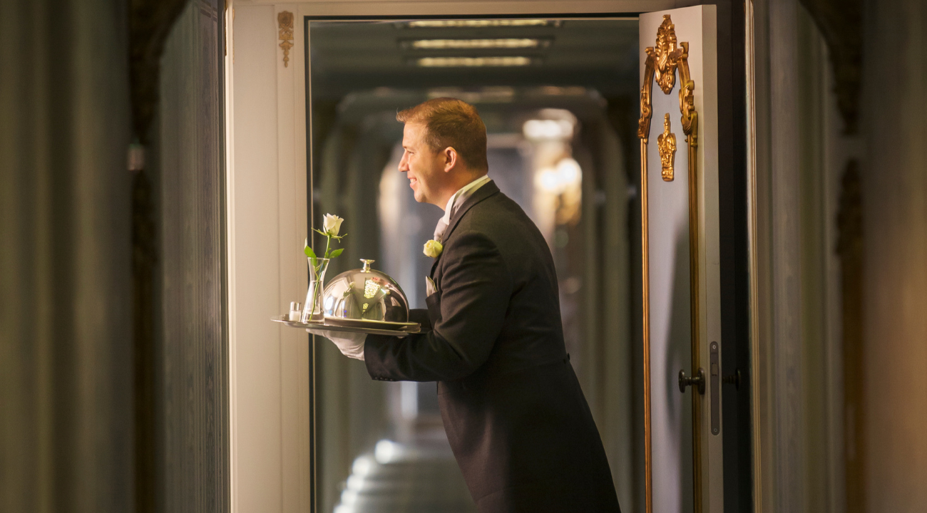 A professional butler in formal attire providing personalised service to a guest onboard the S.S. Joie de Vivre.